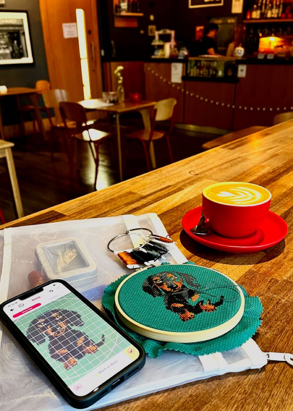 A compact zippered project bag sitting open on a wooden cafe counter, containing a mini cross stitch hoop, smartphone with pattern app, and colourful floss beside a coffee.