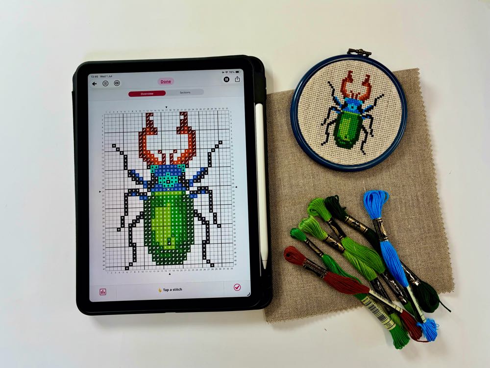 A cozy modern cross stitch setup featuring a colourful embroidery hoop next to a tablet showing a digital pattern chart.