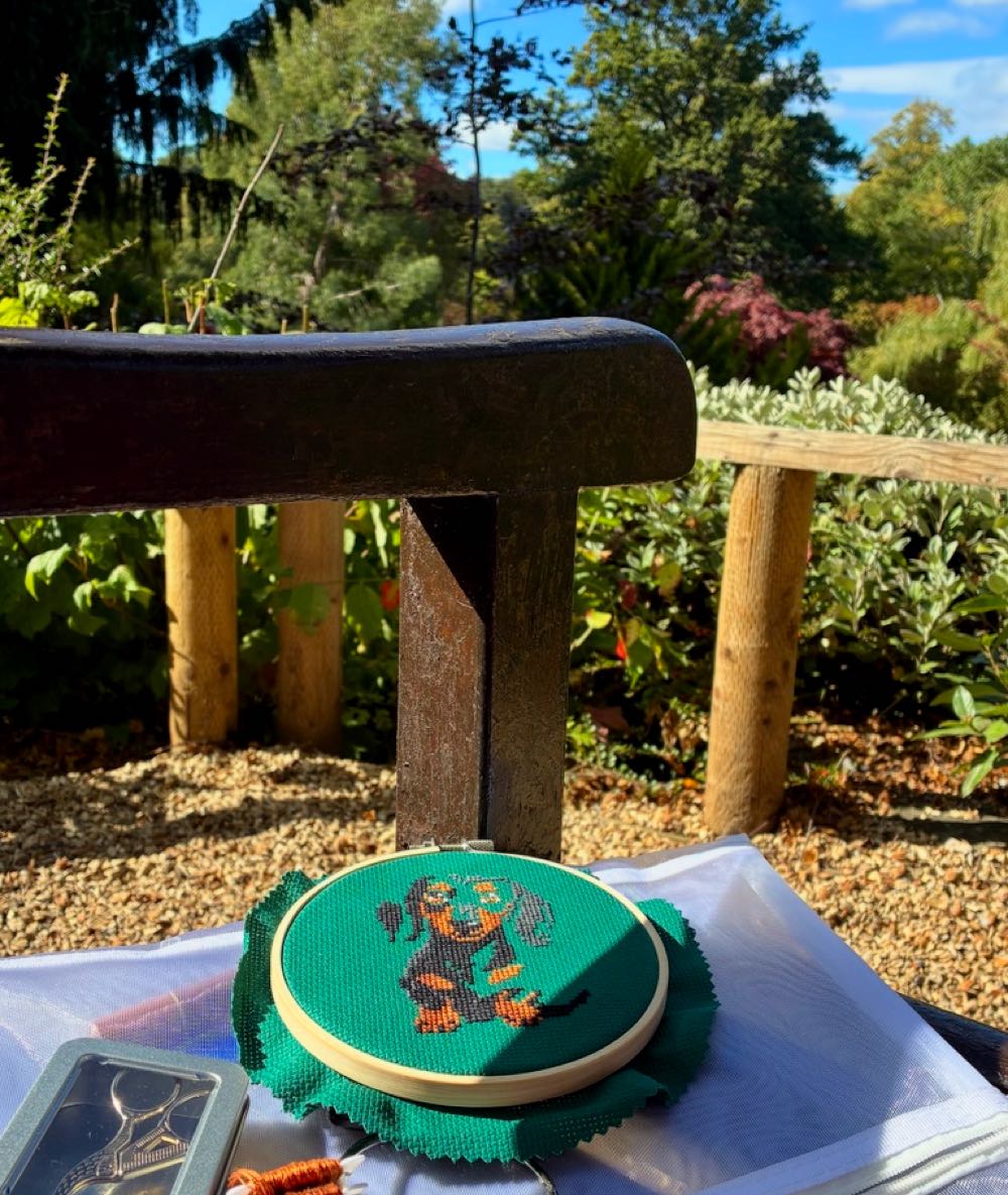 A pocket-sized cross stitch hoop and clear organiser bag resting on a wooden park bench on a sunny day with blue sky overhead.