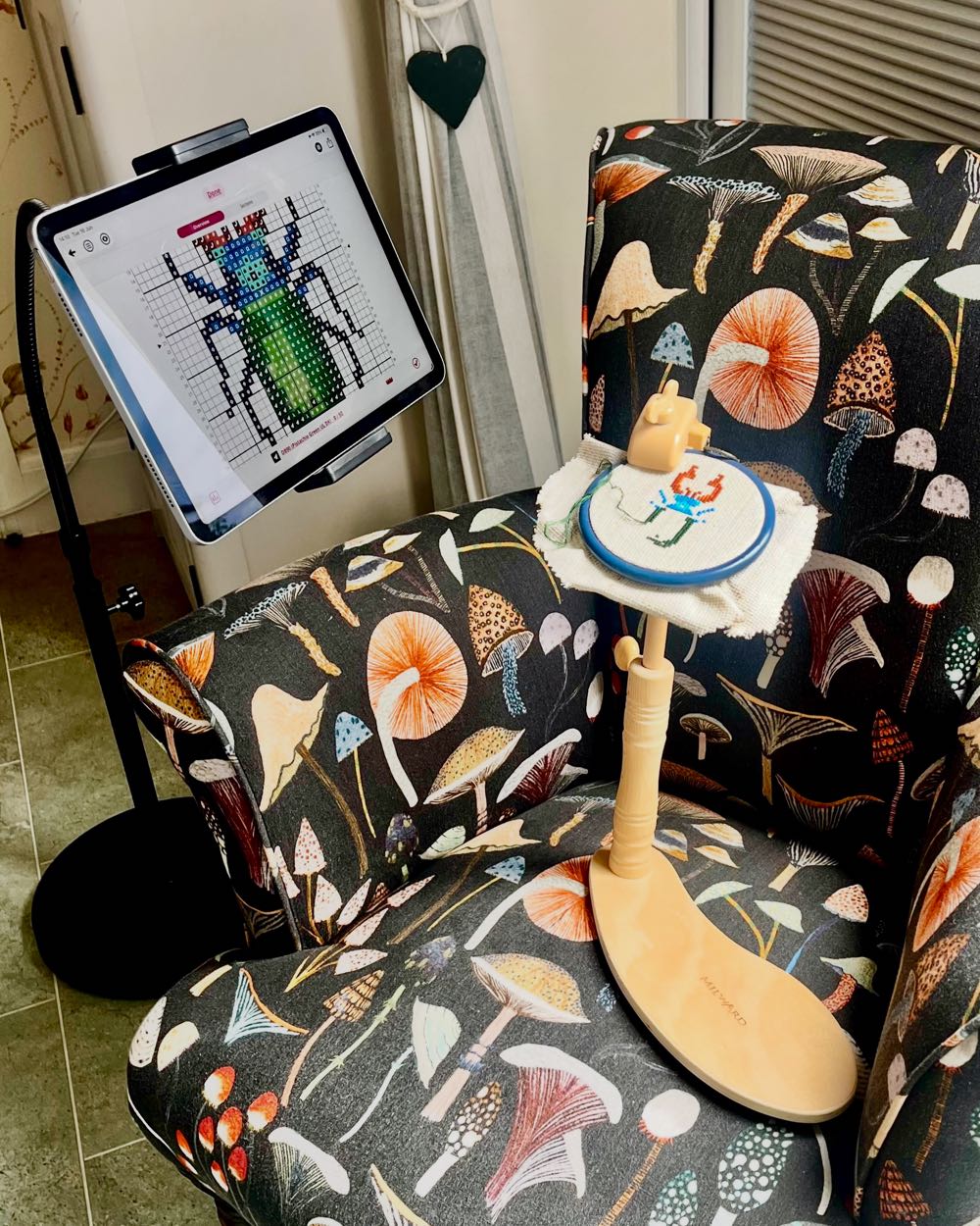 Close up of a tablet held by a goose-neck stand displaying a zoomed-in digital cross stitch chart next to a hoop.