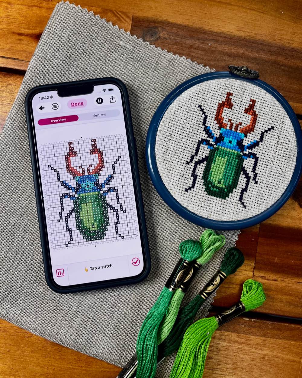 A work in progress cross stitch project resting on a table next to a smartphone showing the track progress screen in the Stitchly app.